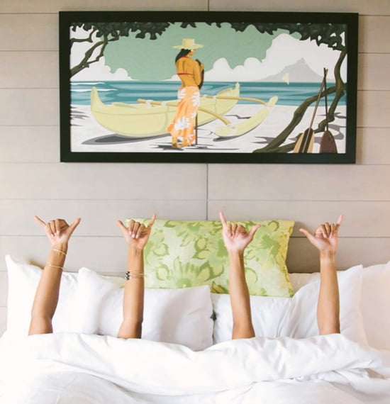 Hands reaching out of a bed making the "hang loose" sign, with an illustration hanging on the wall above them of a woman standing on a beach next to a canoe