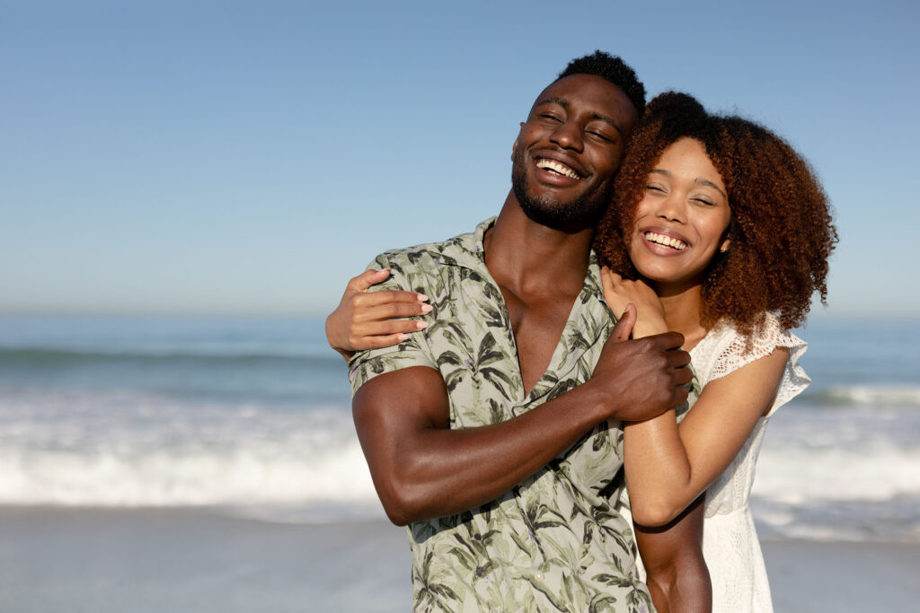 A mixed race couple enjoying free time on beach on a sunny day together, holding each other with sun shining on their faces.