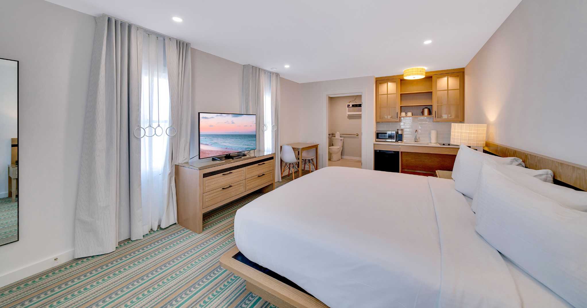 Large king platform bed tv and kitchentte with light wooden accents in the ADA Accessible Hotel Room with Roll in Shower and Kitchenette at Mahalo Cape May beachfront hotel in Cape May, NJ
