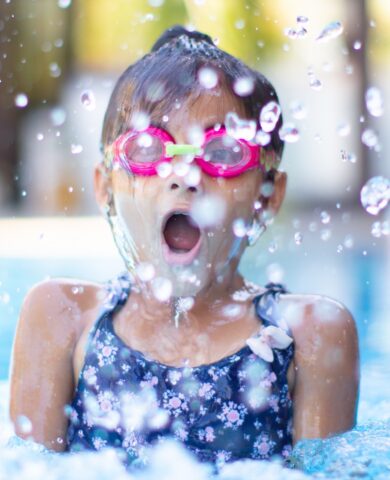 Young girl in goggles gasping in pool with water splashed around her